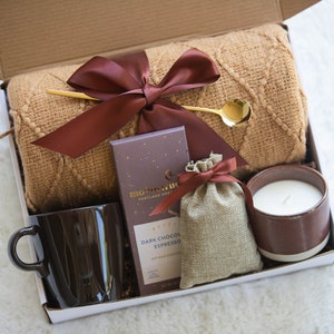 K&ouml;nnte beinhalten: Geschenkbox gef&uuml;llt mit einer braunen Tasse, einer Kerze, einem kleinen Beutel, einer Schokoladentafel, einem goldenen L&ouml;ffel und einer braunen Decke. Die Artikel sind in einer wei&szlig;en Box mit braunem Band angeordnet.
