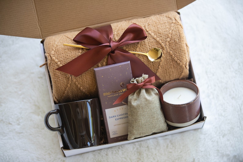 K&ouml;nnte beinhalten: Geschenkbox gef&uuml;llt mit einer braunen Tasse, einer Kerze, einem kleinen Beutel, einer Schokoladentafel, einem goldenen L&ouml;ffel und einer braunen Decke. Die Artikel sind in einer wei&szlig;en Box mit braunem Band angeordnet.