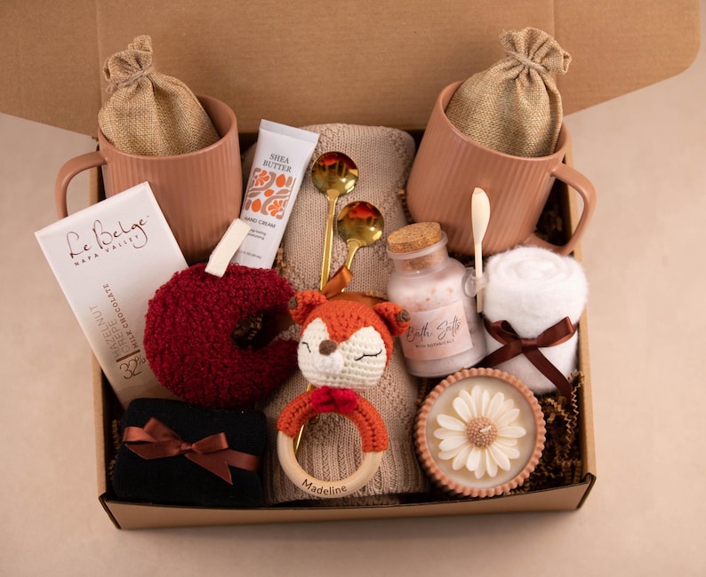May include: A gift box filled with self-care items. Includes two pink mugs with burlap bags, a bar of chocolate, a red apple-shaped item, a tube of hand cream, gold spoons, a crocheted fox, a wooden teething ring, bath salts, a rolled towel, and a candle.