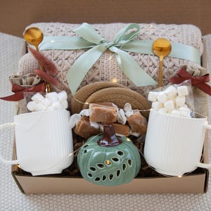 Puede incluir: Una caja de regalo llena de una manta beige de punto trenzado, dos tazas blancas, cucharas doradas, malvaviscos, caramelos y un candelabro verde con forma de calabaza. Una cinta verde claro adorna la manta.