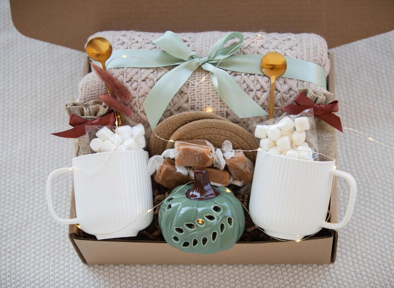 Puede incluir: Una caja de regalo llena de una manta beige de punto trenzado, dos tazas blancas, cucharas doradas, malvaviscos, caramelos y un candelabro verde con forma de calabaza. Una cinta verde claro adorna la manta.