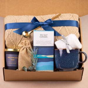 K&ouml;nnte beinhalten: Eine Geschenkbox mit einer beigefarbenen Strickdecke, die mit einem marineblauen Band gebunden ist, einer blauen Keramiktasse, einer Kerze, einem kleinen Beutel, einem St&uuml;ck Seife und einem goldenen L&ouml;ffel. Auf der Kerze steht "Birchwood Tide". Auf der Seife steht "Sea Salt Caramel".