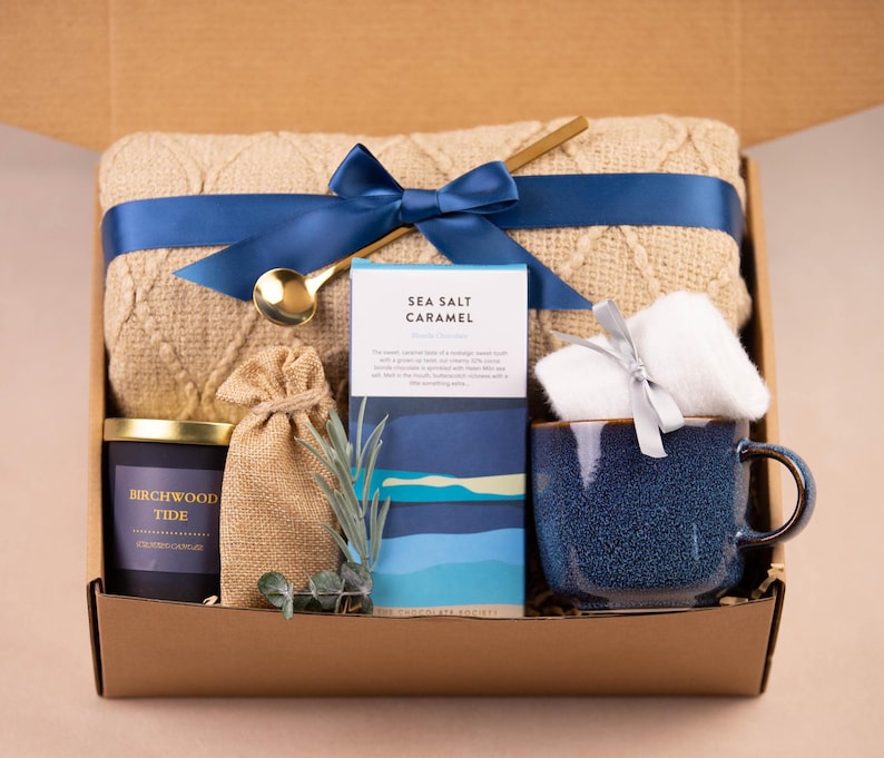 K&ouml;nnte beinhalten: Eine Geschenkbox mit einer beigefarbenen Strickdecke, die mit einem marineblauen Band gebunden ist, einer blauen Keramiktasse, einer Kerze, einem kleinen Beutel, einem St&uuml;ck Seife und einem goldenen L&ouml;ffel. Auf der Kerze steht "Birchwood Tide". Auf der Seife steht "Sea Salt Caramel".
