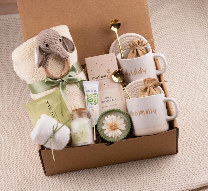 May include: A gift box filled with baby items and self-care products. Includes a cream-colored blanket, a wooden elephant teether, two mugs that say "daddy" and "mommy", and other items. The box is tied with a green ribbon.