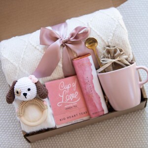 May include: A gift box filled with items for a new parent. The box contains a white blanket with a pink ribbon, a pink mug, a small bag of tea, a bar of chocolate, a wooden rattle with a crocheted dog, and a gold spoon.