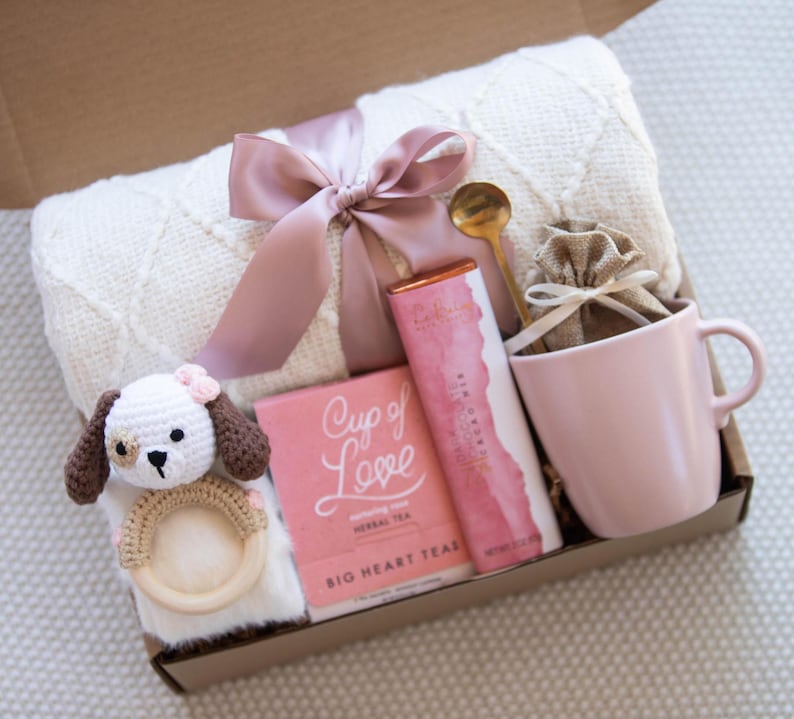 May include: A gift box filled with items for a new parent. The box contains a white blanket with a pink ribbon, a pink mug, a small bag of tea, a bar of chocolate, a wooden rattle with a crocheted dog, and a gold spoon.