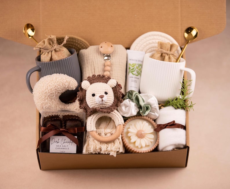 May include: A gift box filled with baby items and self-care products. Includes a crochet lion rattle, a sheep-shaped plush, mugs, a pacifier, a bar of soap, a hand cream, chocolates, and a small towel. The box is brown and has gold spoons attached.