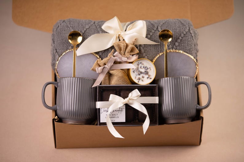 May include: A gift box with two gray ribbed mugs, gold spoons, coasters, and a gray blanket. The box also contains a small candle, a burlap bag, and a box of dark chocolate sea salt caramels, all tied with white ribbons.