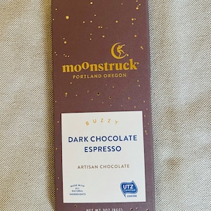 May include: A dark brown chocolate bar with gold foil lettering that reads "moonstruck" and "PORTLAND OREGON". The bar is labeled "BUZZY DARK CHOCOLATE ESPRESSO ARTISAN CHOCOLATE" and has a gold star pattern. The bar is 3 ounces (85g).