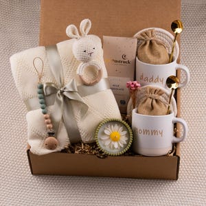 May include: A gift box filled with baby items and gifts for parents. The box contains a cream-colored baby blanket, a crochet bunny rattle, a pacifier, two white mugs with "daddy" and "mommy" printed on them, and other items.