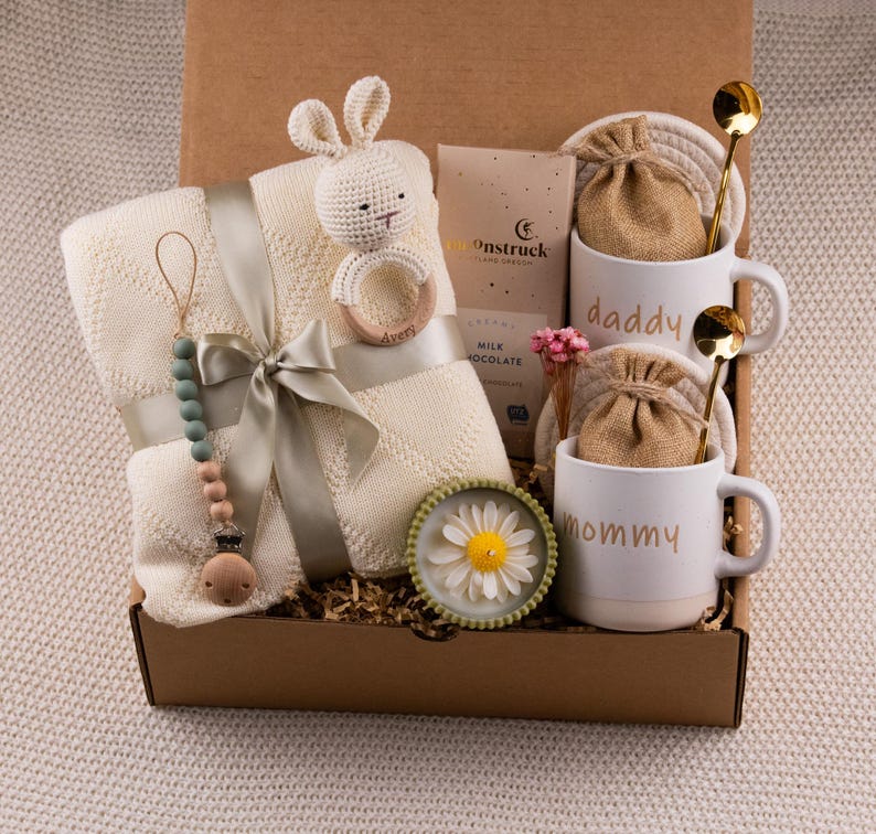 May include: A gift box filled with baby items and gifts for parents. The box contains a cream-colored baby blanket, a crochet bunny rattle, a pacifier, two white mugs with "daddy" and "mommy" printed on them, and other items.