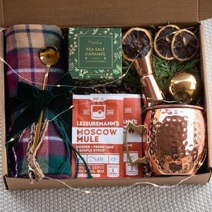 May include: A gift box filled with items for making a Moscow Mule cocktail. The box includes a copper mug, a bottle of Moscow Mule mix, a jar of sea salt caramel, dried orange slices, a plaid throw blanket, and a gold-coloured cocktail stirrer.