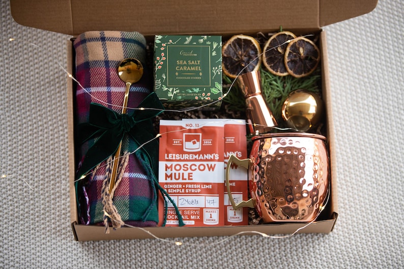 May include: A gift box filled with items for making a Moscow Mule cocktail. The box includes a copper mug, a bottle of Moscow Mule mix, a jar of sea salt caramel, dried orange slices, a plaid throw blanket, and a gold-coloured cocktail stirrer.