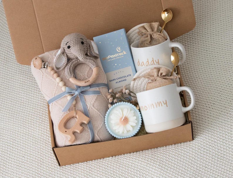 May include: A gift box filled with baby items. Includes a beige blanket, a crochet elephant rattle, a wooden teether, a blue ribbon, a bar of dark chocolate almond, two white mugs with "daddy" and "mommy" in gold lettering, a candle, and two gold spoons.