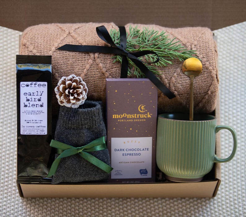 May include: A gift box filled with cosy items for a relaxing evening. It includes a brown knitted throw blanket, a mug, a bag of coffee labelled "coffee early bird blend", a bar of dark chocolate espresso, and a pair of grey socks.