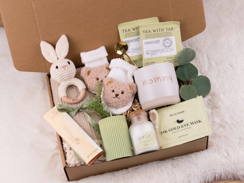 May include: A gift box filled with baby items, including a crochet bunny rattle, bear-faced socks, a mug with "mommy" printed on it, tea packets, a candle, and a jar of bath salts. The box is lined with beige fabric.