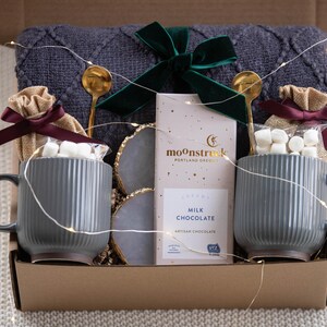 May include: A gift box with a cozy theme. It contains two gray mugs with marshmallows, gold spoons, a bar of "Milk Chocolate", and a dark green bow. The box also includes a gray blanket and decorative lights.