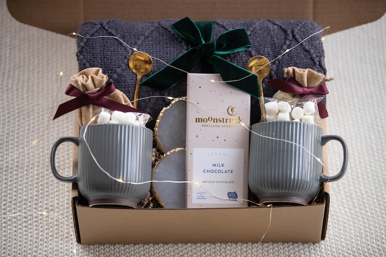 May include: A gift box with a cozy theme. It contains two gray mugs with marshmallows, gold spoons, a bar of "Milk Chocolate", and a dark green bow. The box also includes a gray blanket and decorative lights.