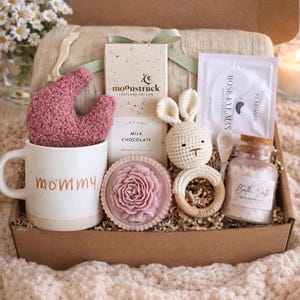 May include: A gift box filled with self-care items. Includes a white mug with "mommy" in gold lettering, a pink sock, a bar of chocolate, a crochet bunny, a wooden teether, bath salts, and a rose eye mask.