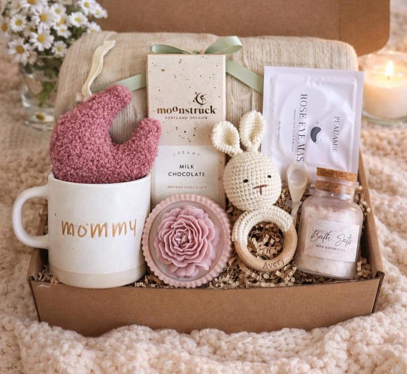 May include: A gift box filled with self-care items. Includes a white mug with "mommy" in gold lettering, a pink sock, a bar of chocolate, a crochet bunny, a wooden teether, bath salts, and a rose eye mask.