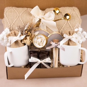 May include: A gift box filled with a beige knitted blanket, two white ribbed mugs, marshmallows, chocolates, and gold spoons. The box is tied with white ribbons and includes decorative elements.