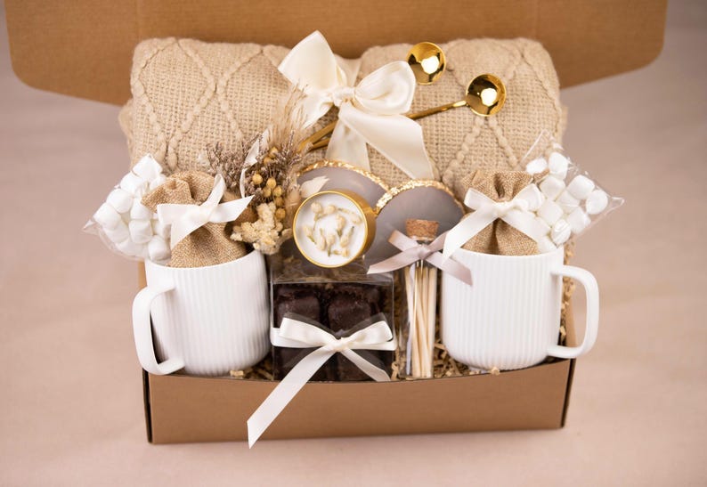 May include: A gift box filled with a beige knitted blanket, two white ribbed mugs, marshmallows, chocolates, and gold spoons. The box is tied with white ribbons and includes decorative elements.