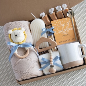 May include: A gift box filled with baby items, including a beige blanket, a lion rattle, a candle, tea, a mug, and caramel candies. The box is tied with blue ribbons, creating a thoughtful baby shower gift.