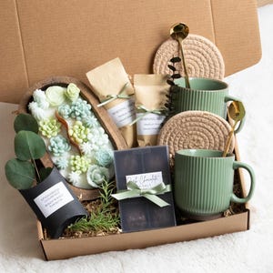 Puede incluir: Una caja de regalo llena de varios art&iacute;culos. Incluye dos tazas verdes, cucharas doradas y posavasos tejidos. Tambi&eacute;n hay un cuenco de madera con jabones en forma de suculentas, dos bolsas de caf&eacute; y una tableta de chocolate negro.