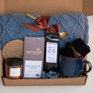 May include: A gift box filled with cosy items, including a blue knitted throw blanket, a mug, a candle, a bar of dark chocolate espresso, a bag of coffee labelled "coffee early bird blend", and a pair of black socks.