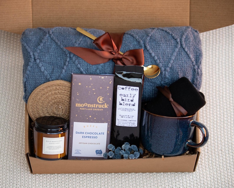 May include: A gift box filled with cosy items, including a blue knitted throw blanket, a mug, a candle, a bar of dark chocolate espresso, a bag of coffee labelled "coffee early bird blend", and a pair of black socks.