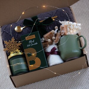May include: A gift box filled with cosy items for a relaxing evening. It includes a green mug, a candle, a blanket, hot chocolate, marshmallows, and caramel sweets. The box is decorated with a snowflake and fairy lights.