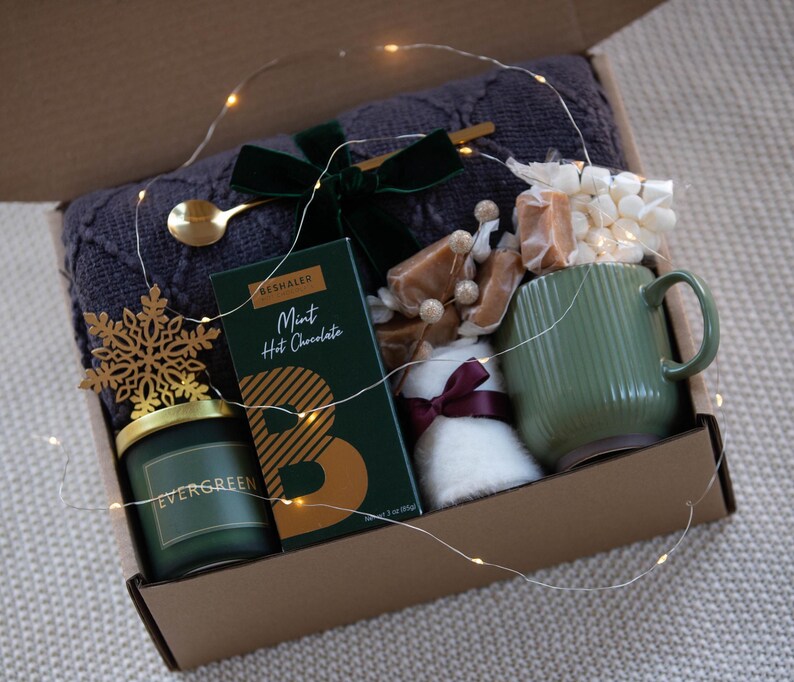 May include: A gift box filled with cosy items for a relaxing evening. It includes a green mug, a candle, a blanket, hot chocolate, marshmallows, and caramel sweets. The box is decorated with a snowflake and fairy lights.