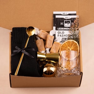 May include: A brown cardboard box filled with a gift set for making an Old Fashioned cocktail. The set includes a black knitted sock, a gold bar spoon, a gold jigger, a gold-coloured ice ball mould, a glass of ice, a dried orange slice, and a bag of caramel sweets. The box also includes a packet of cocktail mix labelled "Leisuremaker Old Fashioned".