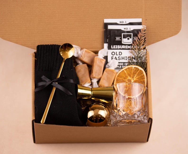 May include: A brown cardboard box filled with a gift set for making an Old Fashioned cocktail. The set includes a black knitted sock, a gold bar spoon, a gold jigger, a gold-coloured ice ball mould, a glass of ice, a dried orange slice, and a bag of caramel sweets. The box also includes a packet of cocktail mix labelled "Leisuremaker Old Fashioned".