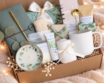 May include: A gift box containing a sage green notebook with a gold pen, a white ribbed mug with a gold spoon, a bar of Moonstruck milk chocolate, a bag of Morning Brew coffee, bath salts, hand cream, a white fuzzy item, and a candle.