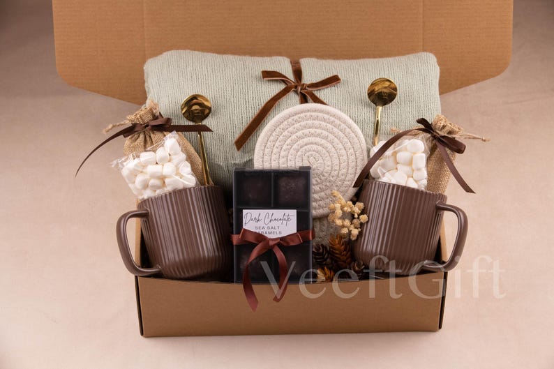 May include: A gift basket filled with a light green blanket, two brown mugs, marshmallows, dark chocolate, and a woven coaster. The basket includes two gold spoons and is tied with a brown ribbon. The dark chocolate label reads "Dark Chocolate Sea Salt Caramels."