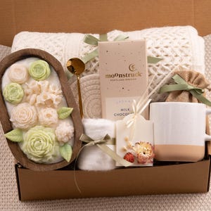 May include: A gift box filled with a variety of items. Includes a wooden bowl with floral soap, a gold spoon, a mug, a chocolate bar, a candle, a coaster, and a small burlap bag tied with a green ribbon.