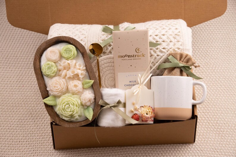 May include: A gift box filled with a variety of items. Includes a wooden bowl with floral soap, a gold spoon, a mug, a chocolate bar, a candle, a coaster, and a small burlap bag tied with a green ribbon.