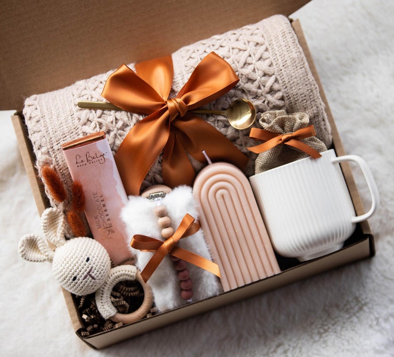 May include: A gift box filled with a variety of items, including a knitted blanket, a candle, a mug, a chocolate bar, a small stuffed bunny, and a wooden rattle. The box is decorated with a brown ribbon.