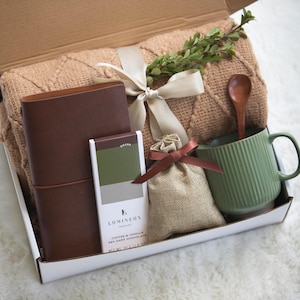 May include: A gift box filled with a brown leather journal, a bar of dark chocolate with the text "Lumineux Coffee & Vanilla 58% Dark Chocolate", a small hessian bag with a brown ribbon, a green ceramic mug with a wooden spoon, and a brown knitted throw blanket.