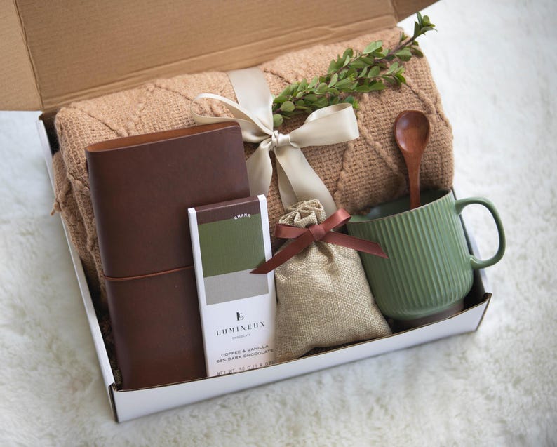May include: A gift box filled with a brown leather journal, a bar of dark chocolate with the text "Lumineux Coffee & Vanilla 58% Dark Chocolate", a small hessian bag with a brown ribbon, a green ceramic mug with a wooden spoon, and a brown knitted throw blanket.