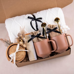 May include: A gift box with a white blanket, two pink mugs, gold spoons, and a woven coaster. The mugs have a vertical ribbed design. The box also contains small burlap bags tied with black ribbons.
