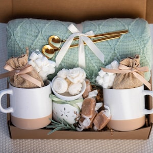 May include: A gift box filled with a light green knitted blanket, two white mugs with tan bottoms, marshmallows, caramels, and gold spoons. The mugs contain burlap bags tied with pink ribbons. A white ribbon and small white bowl are also included.