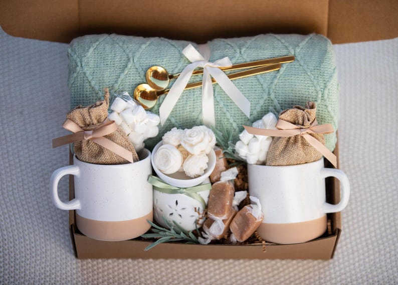 May include: A gift box filled with a light green knitted blanket, two white mugs with tan bottoms, marshmallows, caramels, and gold spoons. The mugs contain burlap bags tied with pink ribbons. A white ribbon and small white bowl are also included.