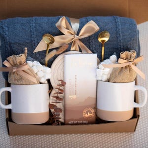 May include: A gift box with a blue blanket, two white mugs with tan bottoms, gold spoons, marshmallows, and a chocolate bar. The box is tied with a gold ribbon and contains a cozy theme.