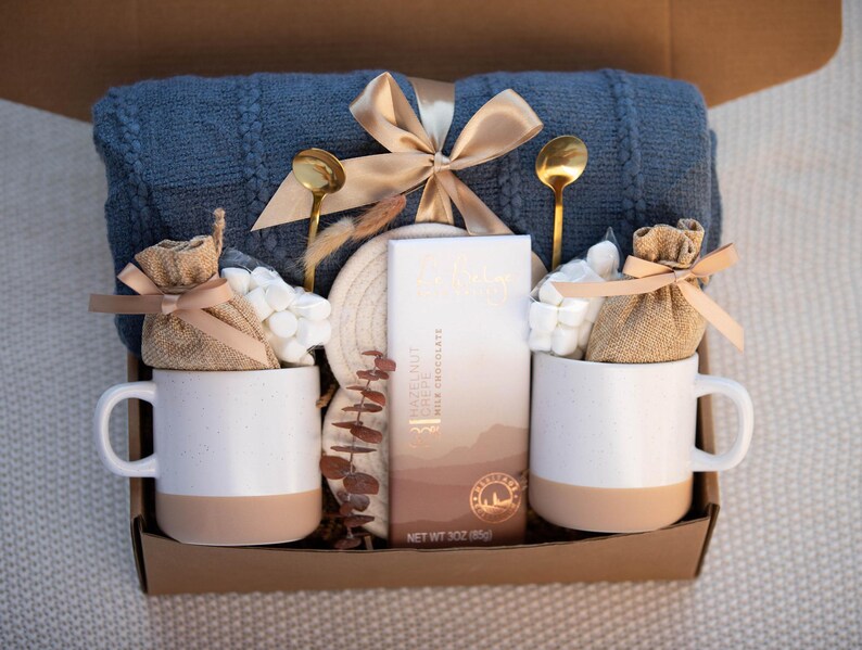 May include: A gift box with a blue blanket, two white mugs with tan bottoms, gold spoons, marshmallows, and a chocolate bar. The box is tied with a gold ribbon and contains a cozy theme.