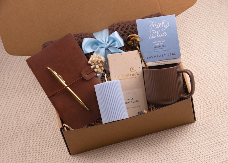 K&ouml;nnte beinhalten: Eine Geschenkbox gef&uuml;llt mit einem braunen Lederjournal und goldenem Stift, einer blauen Kerze, einer Tasse, Tee, Schokolade und einer blauen Schleife. Die Box ist braun und steht auf einer cremefarbenen Oberfl&auml;che.