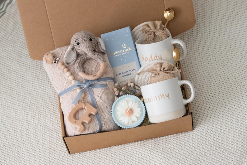 May include: A gift box filled with baby items and treats. Includes a knitted blanket, a crochet elephant rattle, a wooden teether, a bar of dark chocolate almond, two white mugs with "daddy" and "mommy" in gold lettering, and a candle.
