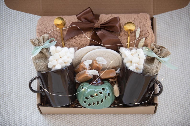 Puede incluir: Una caja de regalo llena con una manta marr&oacute;n, dos tazas marr&oacute;n oscuro, bolsas de malvaviscos, cucharas doradas y caramelos. Una calabaza de cer&aacute;mica verde decorativa y un lazo marr&oacute;n completan el conjunto.