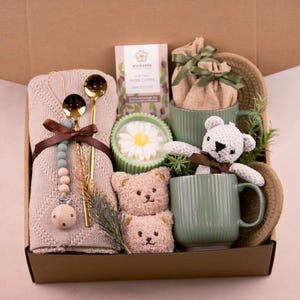 May include: A gift box containing baby-related items. It features a beige blanket, gold spoons, a dummy, two green mugs, a white teddy bear, two small teddy bear faces, a candle, and coffee. The box is brown.